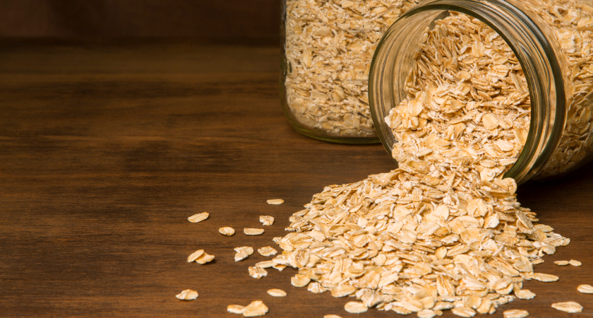 Tight shot of rolled Oats spilling from a glass canning jar onto a rustic dark wool table top with an out-of-focus burlap background.
