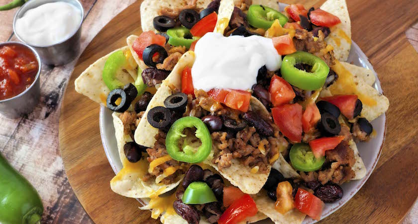 Mexican nacho chips topped with sour cream, ground meat, jalapenos, tomatoes, beans and melted cheese on a wooden paddle board